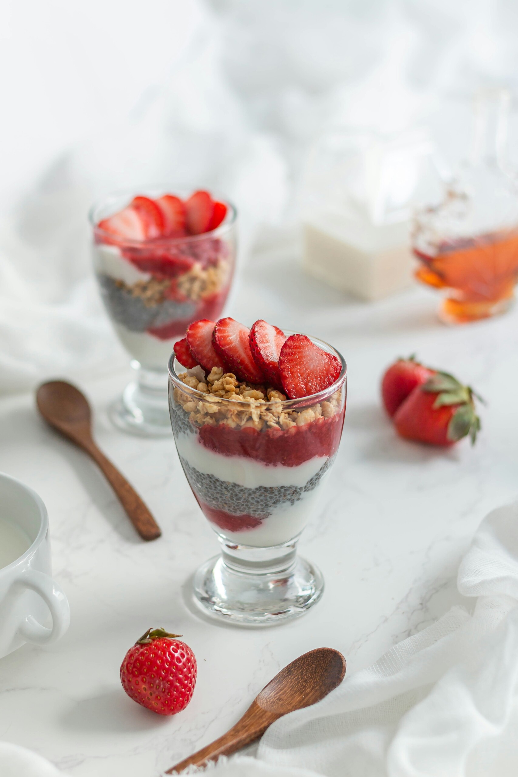 2 yogurt parfaits with strawberries, granola, chia seeds, and yogurt layered in a glass dish. a wooden spoon is beside the 2 dishes. Set on white tablecloth