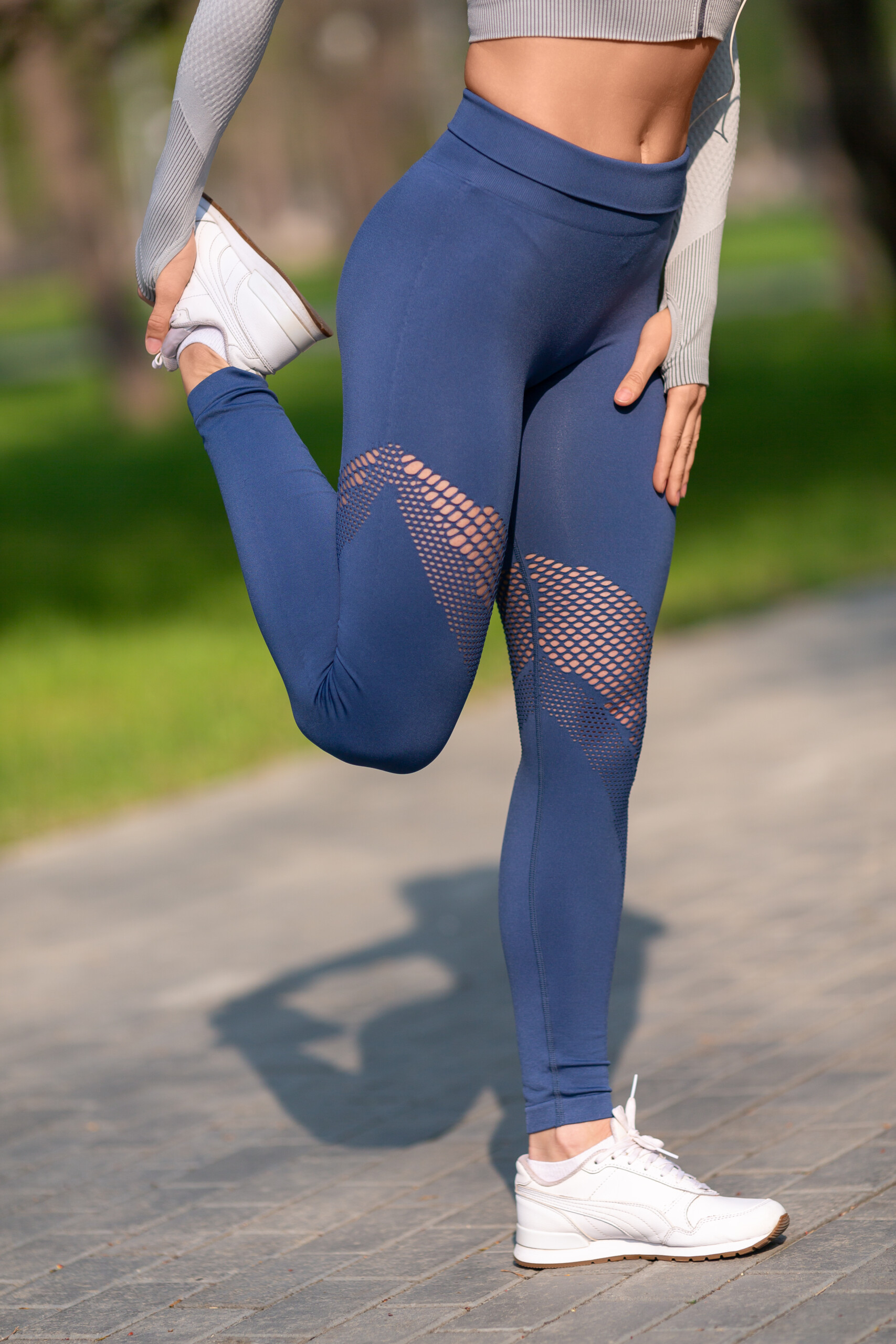 girl with blue leggings, white sneakers stretching her hamstring. She is on pavement with green grass behind her.