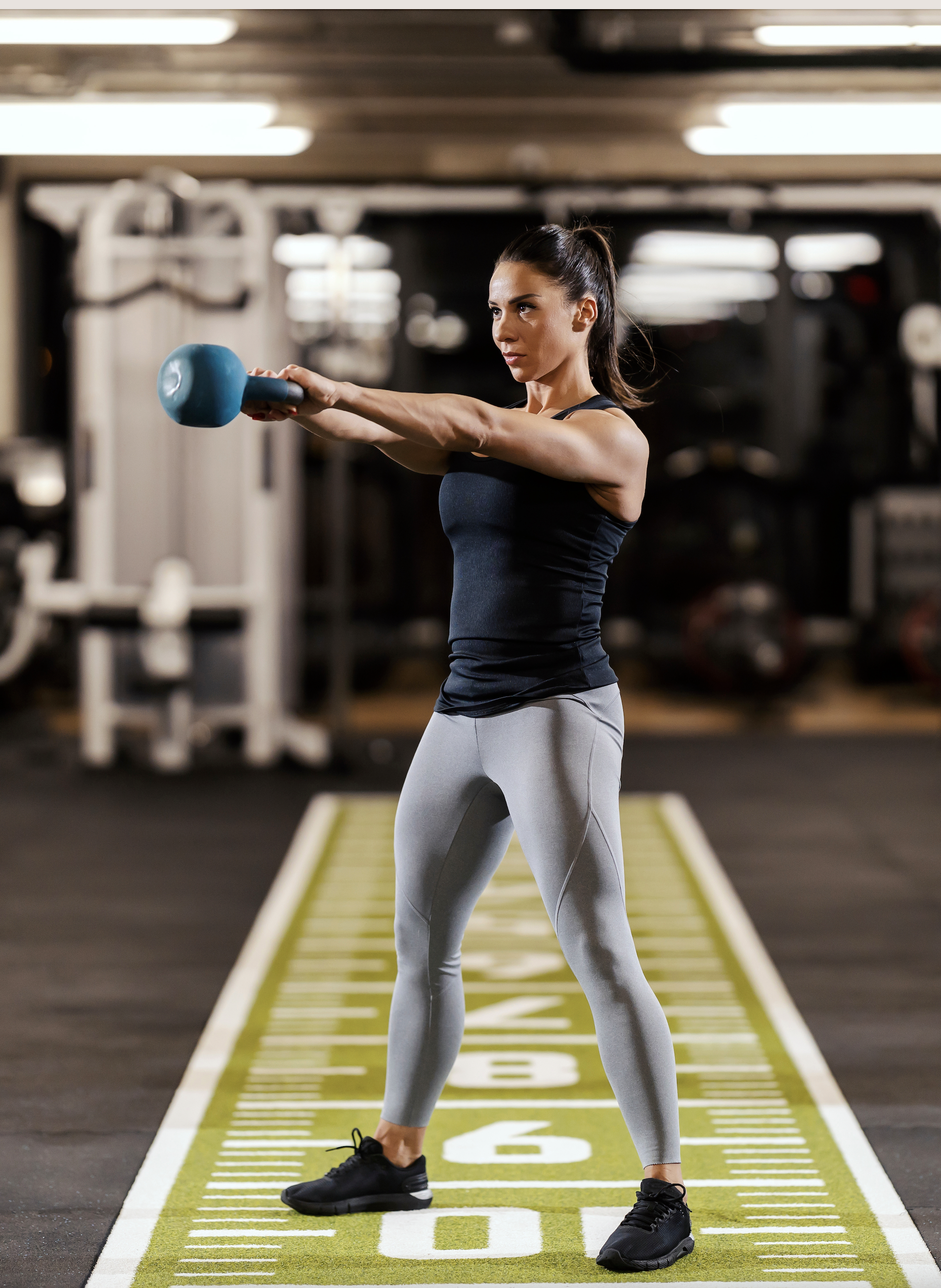 woman in grey leggings and black tank top swinging a blue kettlebell in a gym