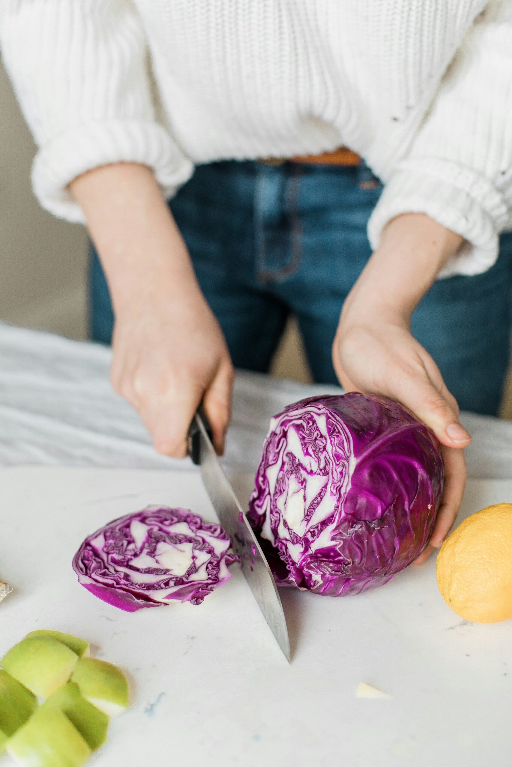 woman with white sweater and jeans cutting a purple cabbage. View is from her waist down