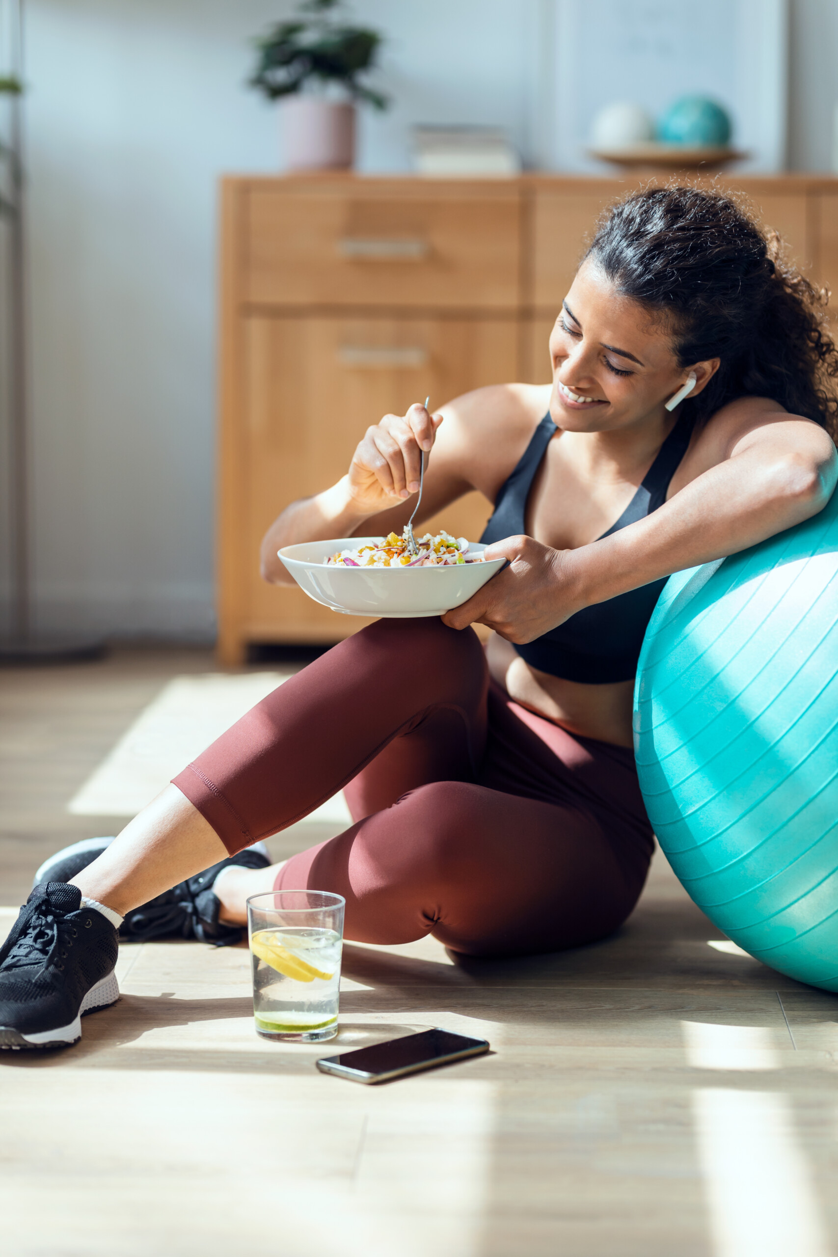 woman in fitness clothes eating a salad leaning on a blue exercise ball with a glass of lemon water beside her. She is indoors.