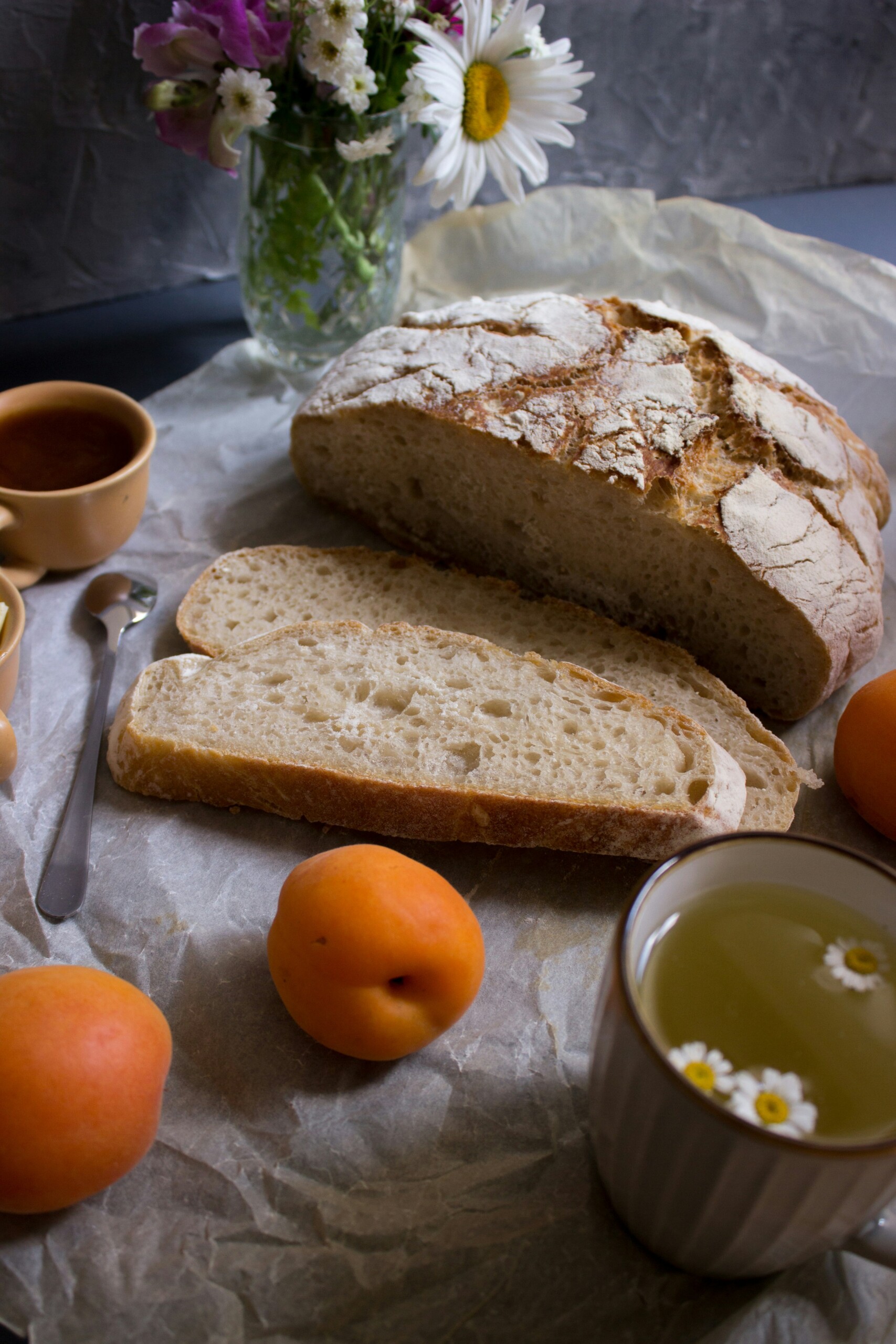 a loaf of sourdough bread with one slice cut, 2 hot tea in cups, 2 oranges, and a spoon on table
