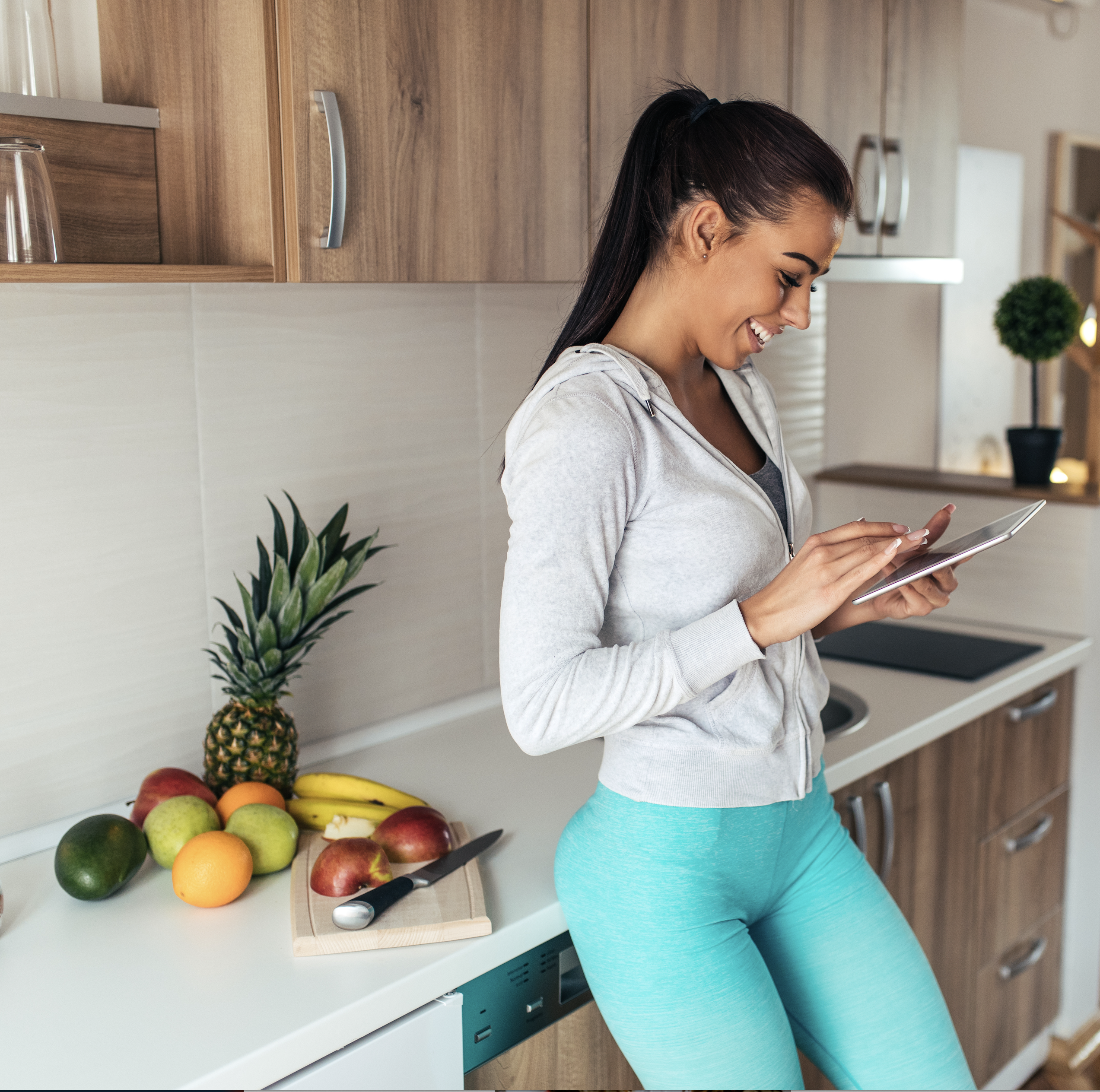 woman with blue leggins and grey hoodie with ipad. She is smiling. She in in a kitchen with fruit behind her. Kitchen has a modern feel.