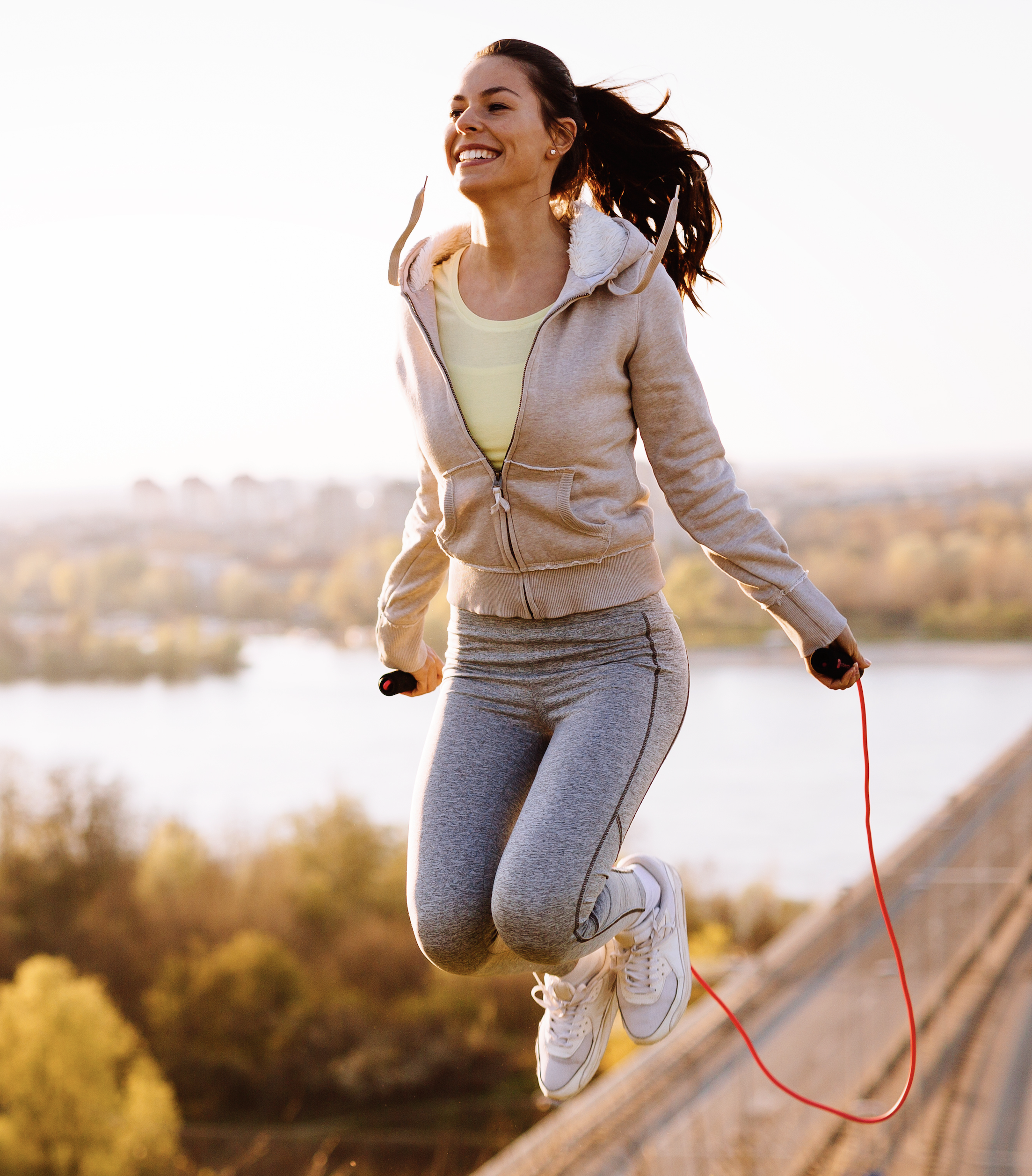 woman in fitness where wearing a beige jacket jumping rope. She has a lake with trees behind her.