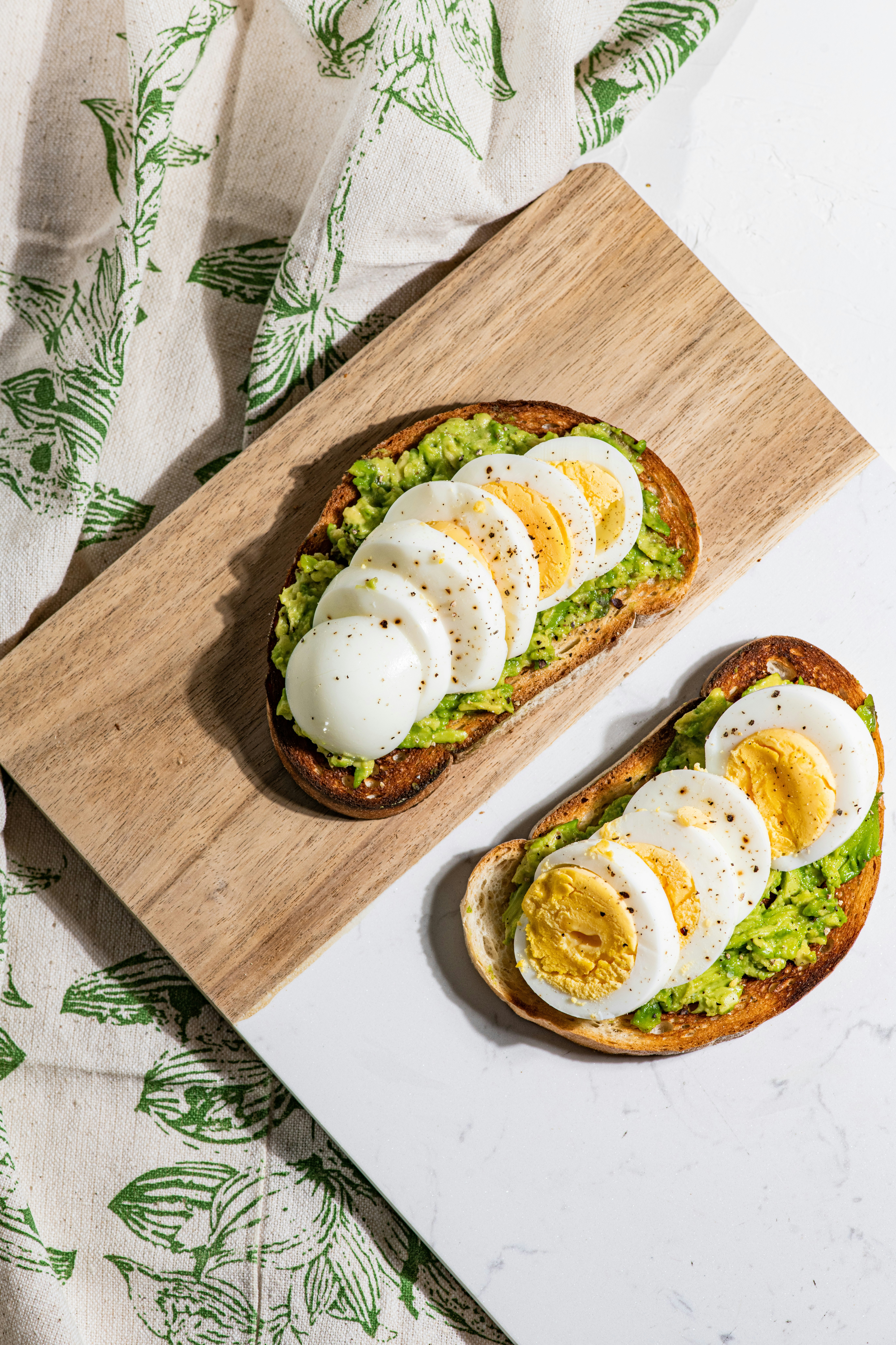 avocado toast with a sliced boiled egg on a cutting board with green toile fabric behind it