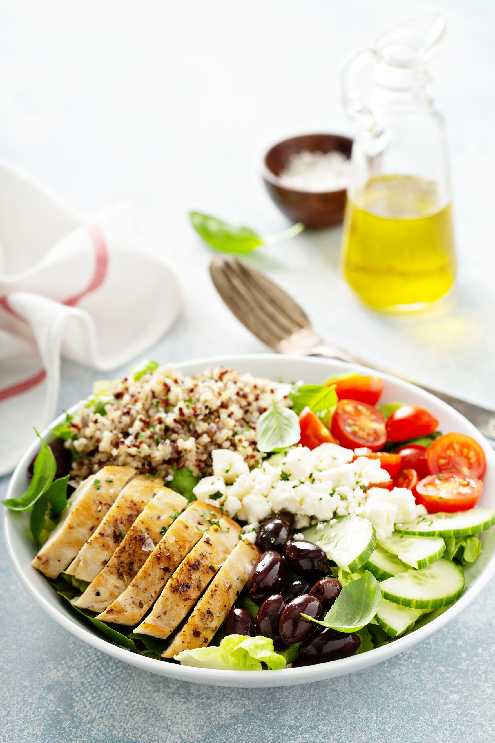 white bowl with chickn breast, quinoa, tomatos cucumbers feta cheese, and olives. a flask of olive oil and bowl of salt on the side