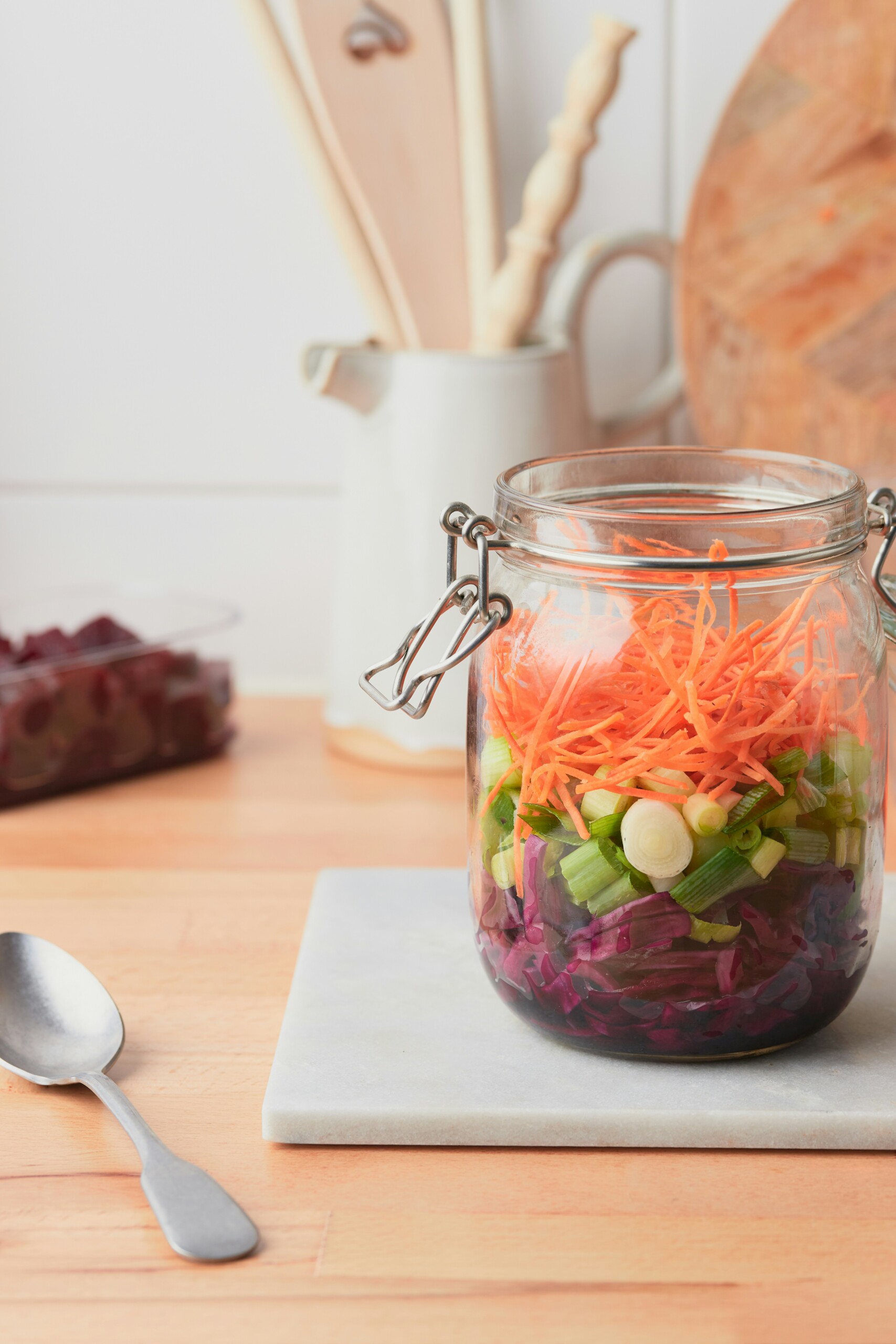 a jar with purple cabbage, oinions and carrots on a cutting board with a s spoon beside it and utensils in the background