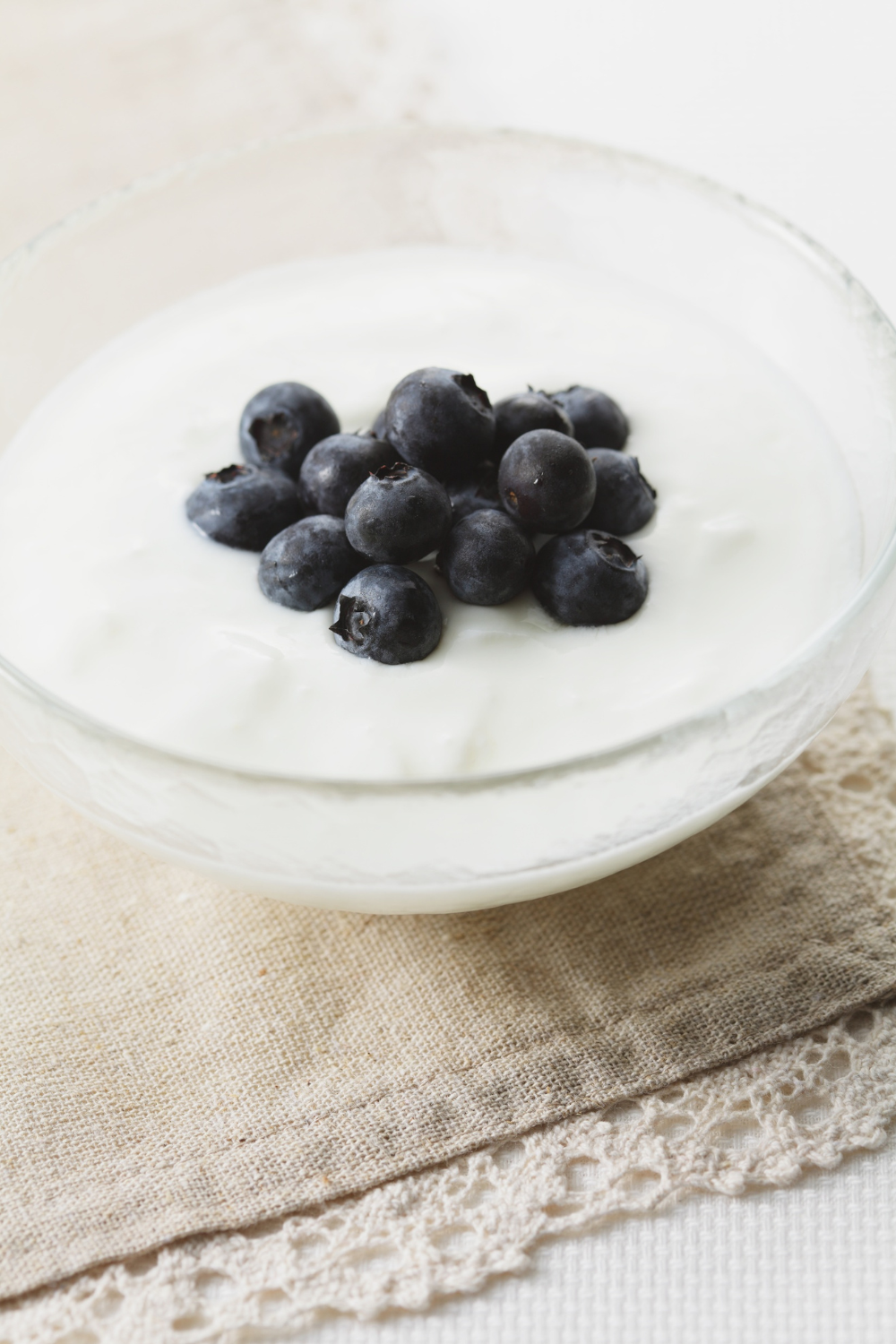 Glass bowl of Greek yogurt topped with fresh blueberries on a neutral linen background